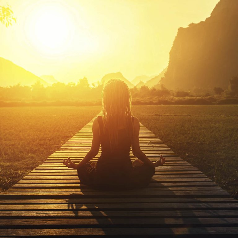 Mujer sentada en posición de meditación, de frente a amanecer